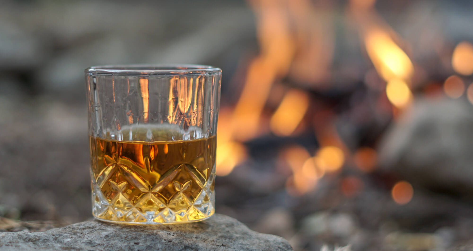 Welche Rum Stile gibt es? Tumbler mit Rum auf Stein vor Lagerfeuer